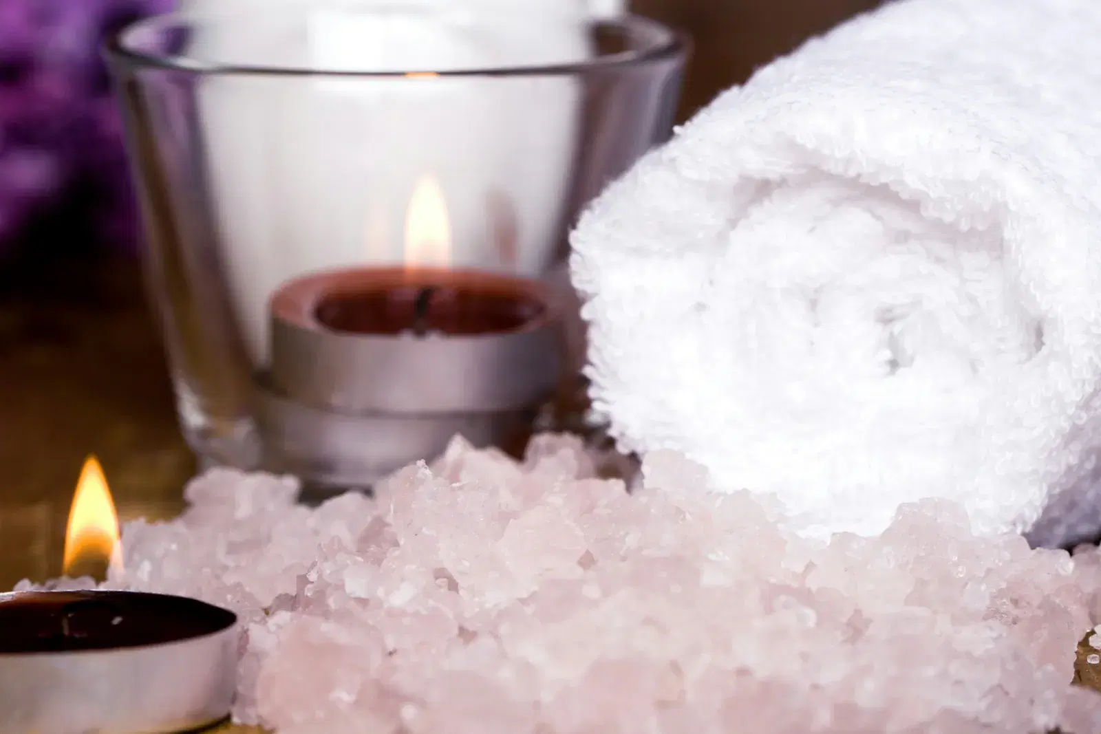 Lit candles beside pink bath salt and a rolled white towel