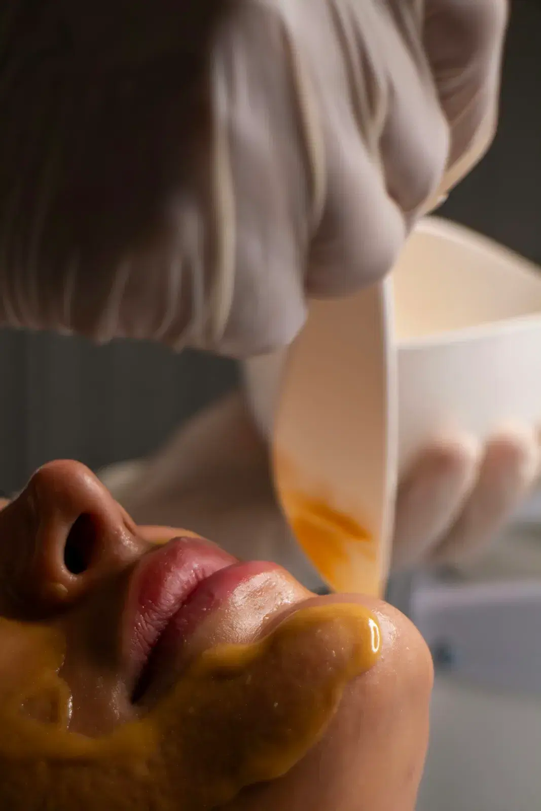A golden facial mask being poured from a bowl onto a person’s chin and cheek