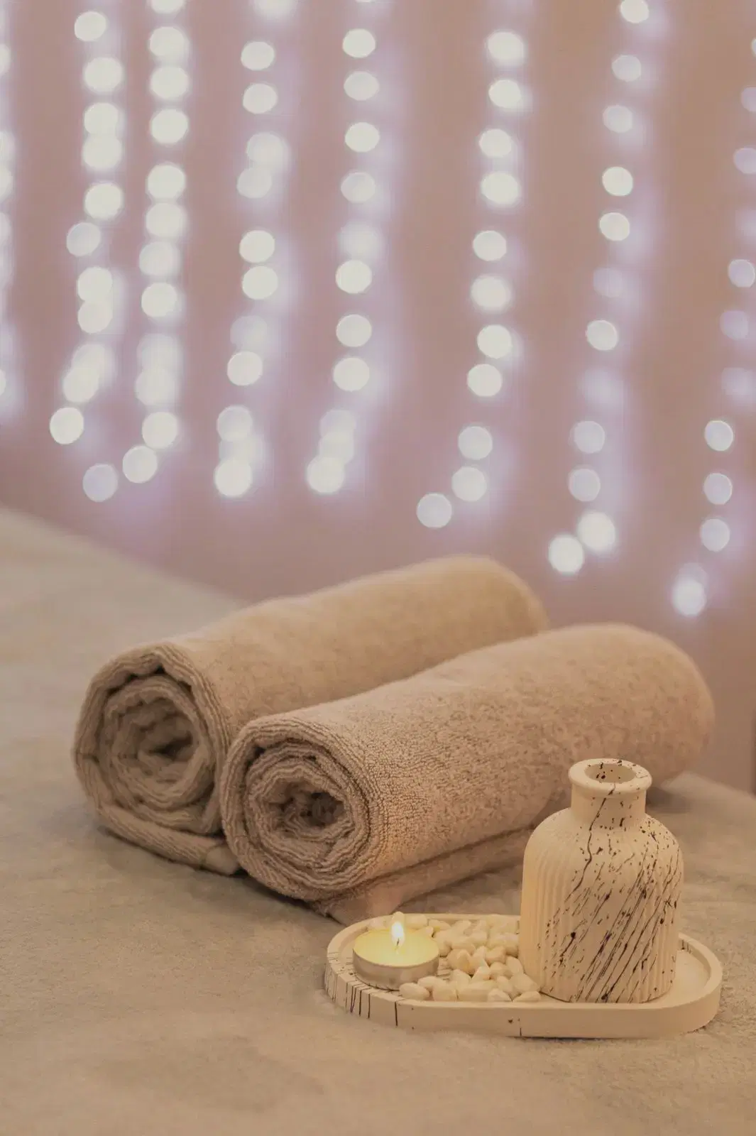 Rolled towels on a treatment bed beside a lit candle, with soft pink lights behind